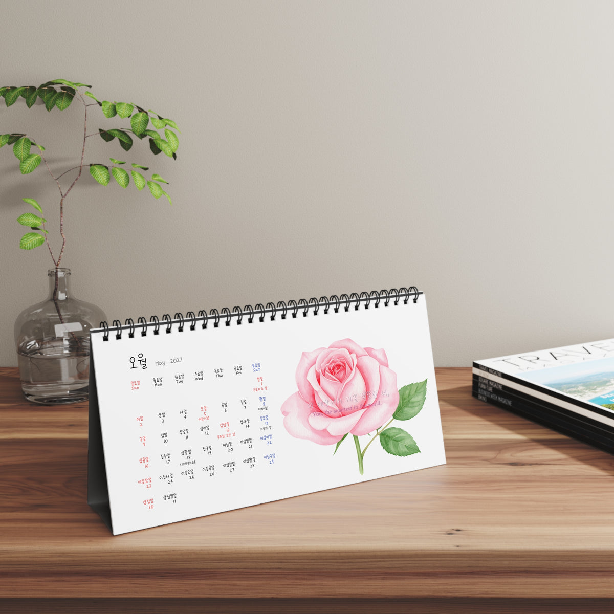 A spiral-bound desk calendar featuring a pink rose illustration on the cover, displayed on a wooden table next to a glass vase with green leaves and a stack of books against a neutral wall. Numbers in Calendar are written in Korea which is great for Korean language learner. Korean-English bilingual.