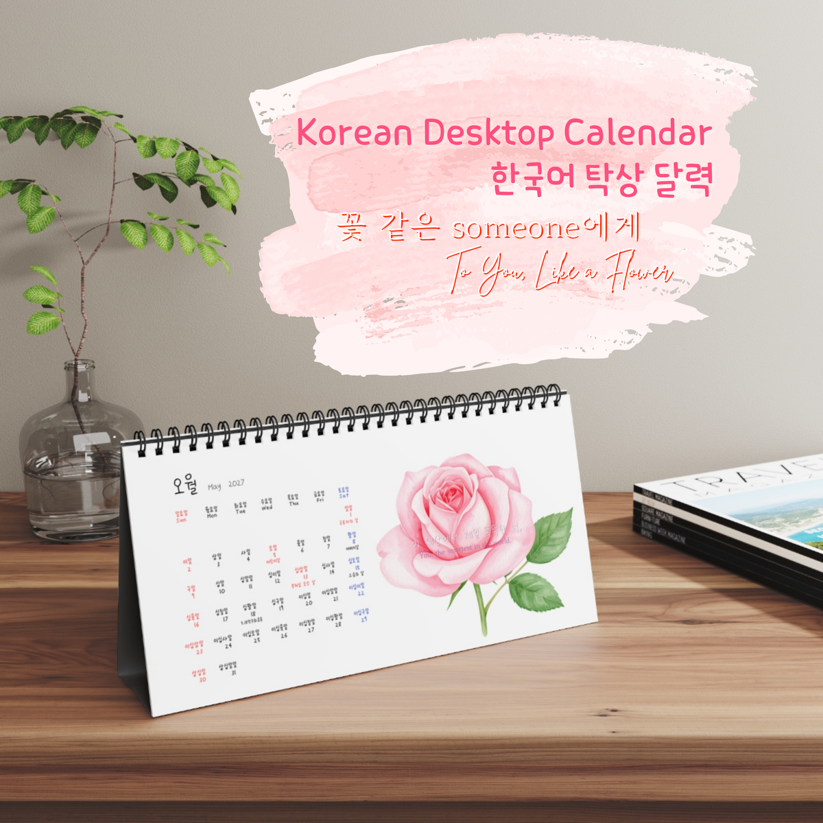 A spiral-bound Korean desktop calendar standing on a wooden table, featuring a pink rose illustration and pink decorative text overlay on a soft pink brushstroke background. The scene includes a glass vase with green stems on the left and a closed book on the right. Korean-English bilingual calendar with numbers and dates written in Korean, designed to help Korean language learners practice numbers naturally.